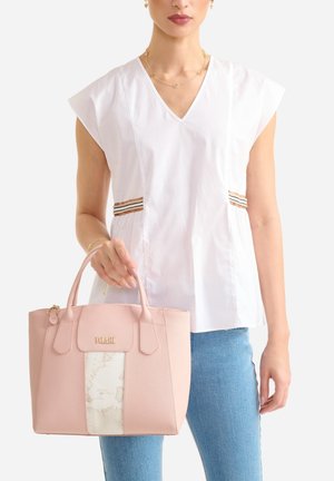 Donna che indossa un top bianco senza maniche con scollo a V e jeans blu, che tiene in mano una borsa rosa con un pannello centrale decorato e un logo dorato.