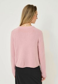 Helles Rosa, weich strukturierter Pullover mit langen Ärmeln und abgerundetem Saum, über einem schwarzen Kleidungsstück getragen. Rückansicht mit minimalen Designdetails.