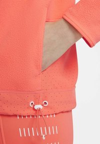 Top de ropa deportiva en color coral con una parte superior de felpa texturizada y una sección inferior lisa con puntos, que cuenta con un cordón para ajustar.