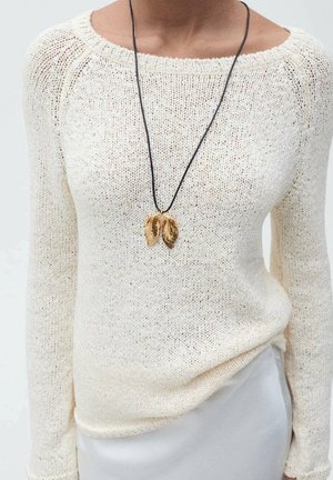 Personne portant un pull en maille crème et un pantalon blanc avec un collier en cordon noir orné de deux pendentifs en forme de feuilles dorées.