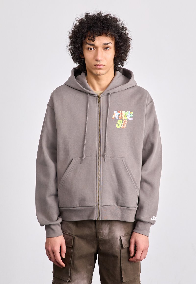 Gray zip-up hoodie made of cotton blend with a front pocket and multicolored "NIKE SB" logo. Features a drawstring hood and ribbed cuffs.