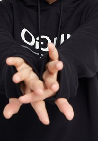 En person som bär en svart huvtröja med vit text, sträcker fram båda händerna med spridda fingrar och delvis döljer texten.