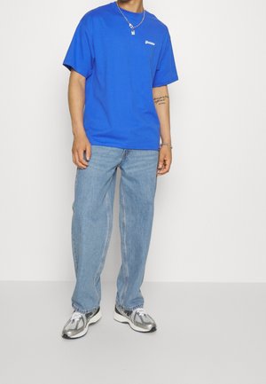 Camiseta de algodón azul con mangas cortas, cuello redondo y un pequeño logo en el lado izquierdo del pecho, combinada con jeans de mezclilla azul claro y zapatillas plateadas.