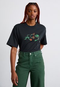 Black cotton t-shirt with short sleeves featuring colorful graphic design. Paired with high-waisted green denim pants.