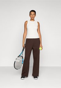Sport tank top in off-white, paired with loose brown pants. Model holds a tennis racket and a green tennis ball, standing against a plain backdrop.