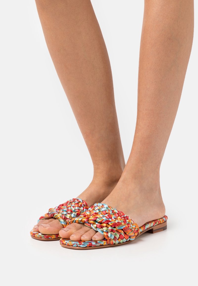 Pieds portant des sandales plates à bout ouvert avec des brides tressées colorées en rouge, orange, bleu et jaune sur fond blanc.