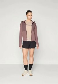 Ung kvinne står iført en mauve zip-up hettegenser, beige T-skjorte, svarte shorts, svarte ankelsokker og hvite joggesko foran en ensfarget bakgrunn.