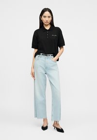 Γυναίκα που φοράει μαύρο πόλο πουκάμισο και ανοιχτό μπλε τζιν με ψηλή μέση και φαρδιά μπατζάκια με ξέφτια στο τελείωμα, συνδυασμένα με μαύρα παπούτσια με μυτερή μύτη και τακούνι kitten.