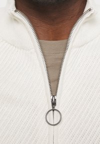 Bílý svetr na zip se zvláštní texturou, který má kovový zip se zaobleným táhlem a béžovou vnitřní vrstvu viditelnou pod ním.