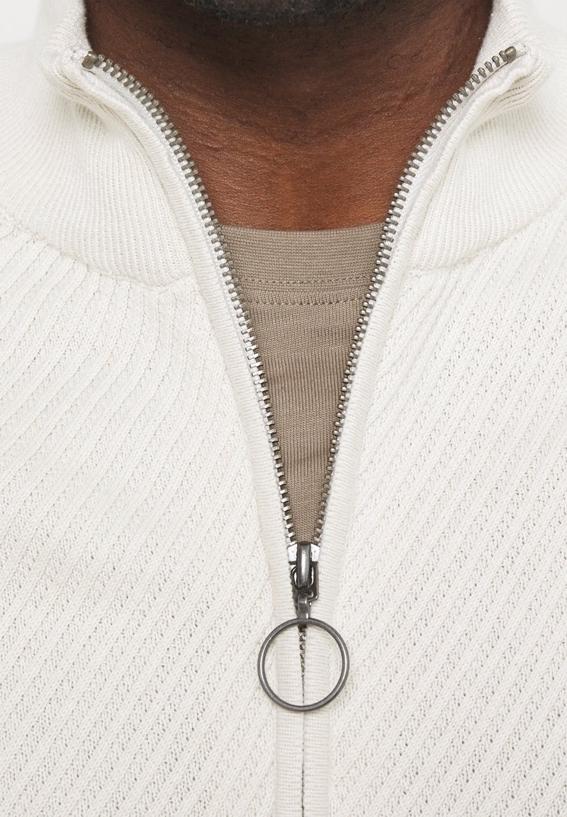 Bílý svetr na zip se zvláštní texturou, který má kovový zip se zaobleným táhlem a béžovou vnitřní vrstvu viditelnou pod ním.