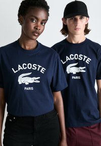 Tricouri din bumbac de culoare albastru marin, cu logo-ul alb Lacoste și o grafică cu crocodil, purtate cu pantaloni scurți negri și bordo, având mâneci scurte.