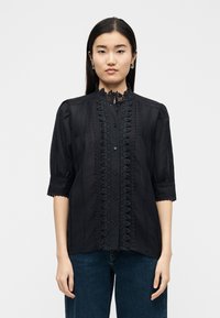 Γυναίκα με μακριά ίσια μαλλιά, φορεμένη με μαύρο μπλουζάκι με δαντέλα στο τελείωμα, κουμπιά και μανίκια μέχρι τον αγκώνα, συνδυασμένο με σκούρο μπλε τζιν.