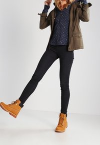 Persoon in tan werklaarzen, zwarte skinny jeans, een marineblauwe blouse en een olijfgroene jas, die één been iets naar voren heft.
