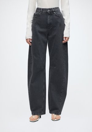 Person trägt schwarze, weit geschnittene High-Waist-Jeans, ein weißes Langarm-Oberteil und weiße Sandalen mit offener Zehenpartie vor einfachem Hintergrund.