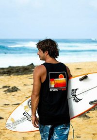 Schwarzes ärmelloses T-Shirt mit retro Grafik, blaue gemusterte Badehose, hält ein weißes Surfbrett mit orangefarbigen Akzenten, am Strand.