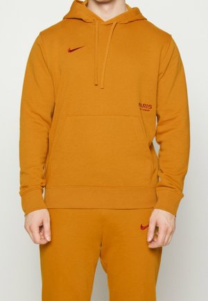 Mężczyzna ubrany w musztardowo-żółtą bluzę z kapturem Paris Saint-Germain Nike oraz pasujące spodnie dresowe, stoi z rękami swobodnie opuszczonymi wzdłuż ciała.