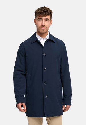 Mann trägt einen marineblauen, geknöpften Mantel über einem weißen Hemd und beigen Hosen, steht vor einem einfachen, hellgrauen Hintergrund.