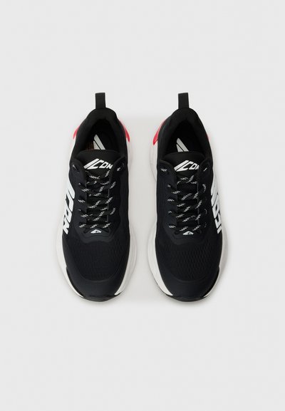 Black athletic shoes with white logo, textured mesh upper, black laces, and red heel accents. Thick white midsole for cushioning.