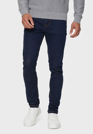 Hombre que lleva vaqueros ajustados azul oscuro, suéter gris y zapatillas blancas, de pie contra un fondo neutro.