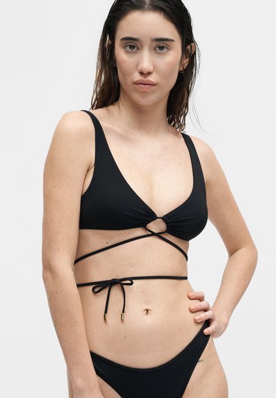 Γυναίκα που φοράει μαύρο μπικίνι με κορδόνια δεμένα σε φιόγκο γύρω από τη μέση της, με μονόχρωμο φόντο.