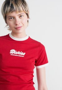 Raudona trumpos rankovės marškinėliai su baltais apykaklės ir rankovių akcentais, su baltu logotipo tekstu "Dickies Workwear Company" priekyje.