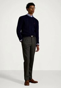 Marinblå stickad tröja över en ljusblå skjorta med krage, grå byxor och bruna läderskor, med runda glasögon och diskret varumärke synligt.