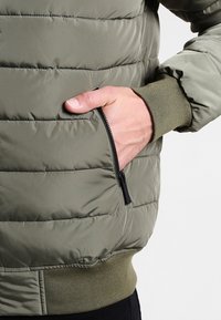 Chaqueta verde acolchada con puños y dobladillo acanalados, bolsillo lateral con cremallera, textura suave y detalle de costura horizontal.