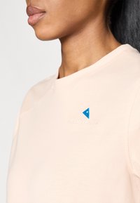 Lyserød t-shirt med rund halsudskæring. Har en blå, trekantet broderet detalje og subtil logo tekst nær skulderen. Blød stofstruktur.