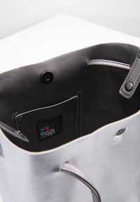 Sac fourre-tout en cuir argenté avec texture lisse, comportant une poche intérieure et une étiquette logo, construit avec des poignées robustes et une fermeture à pression.