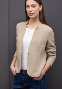 Blazer beige texturé avec un col rond, un seul bouton et des manches longues, porté sur un haut blanc et associé à un jean bleu.