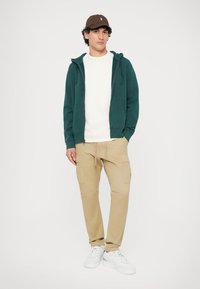Sweatshirt verde com capuz sobre uma camisa branca, combinado com calças cargo bejes e ténis brancos. Um boné castanho adiciona detalhe ao visual.