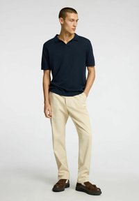 Marinblå polo med kort ärm, ljusbeige byxor och mörka skor. Tyget verkar slätt och lätt. Enkel kragsdesign.