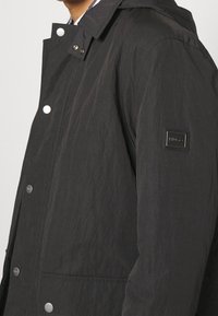 Giacca leggera nera con una finitura testurizzata, bottoni a scatto e una patch con logo sulla manica sinistra. Include un cappuccio e tasche frontali.