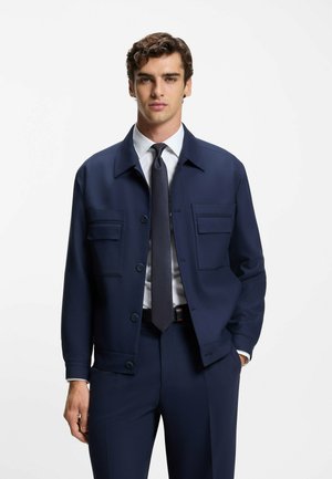 Marineblauwe jas van gestructureerde stof met een kraag, twee zakken aan de voorkant en knoopsluiting, gedragen over een wit overhemd en stropdas.