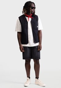 Schwarze ärmellose Weste über einem weißen T-Shirt, kombiniert mit dunklen Jeansshorts und cremefarbenen High-Top-Sneakern. Ein rotes Bandana und Sonnenbrille runden den Look ab.