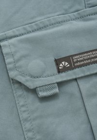 Ljust blått tyg, platt ficka med klaff och etikett som läser "UNDERGROUND DENIM OF NINETEENEIGHTY," som visar upp fin sömnad och textur.