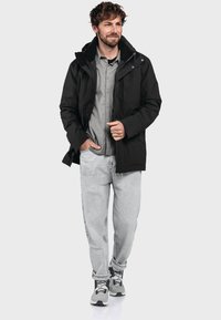 Schwarze Kapuzenjacke mit Reißverschluss, geschichtetes Design, weiche Textur, kombiniert mit einem grauen Shirt, hellen Jeans und grauen Turnschuhen.