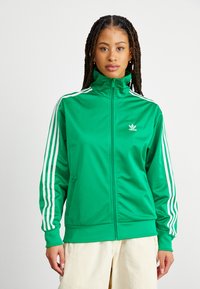 Grön idrottsjacka i slät polyester, med vita ränder längs ärmarna och en broderad logotyp på bröstet. Dragkedja fram, hög krage.