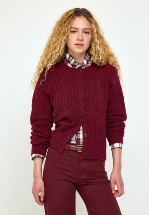 Ung kvinde med langt krøllet blondt hår iført en bordeaux kabelstrikket cardigan over en ternet skjorte og matchende bordeaux bukser.