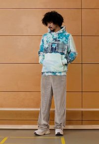 Hanorac cu glugă în model tie-dye albastru-verzui, cu buzunar kangaroo și șnururi albe, asortat cu pantaloni din catifea în culoare gri deschis.