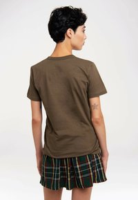 Olivgrünes Baumwoll-T-Shirt mit Rundhalsausschnitt, kurzen Ärmeln und legerer Passform, kombiniert mit einem karierten ausgestellten Rock mit roten, gelben und grünen Streifen.