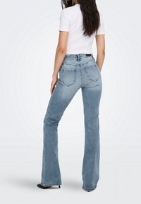 Femme portant un jean évasé bleu clair taille haute et un top blanc à manches courtes, debout dos à la caméra.