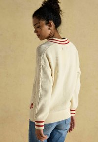 Kreemi sviiter cable-knit musteriga, ribidega punaste triipudega krae ja varrukad ning tikand detail allääre lähedal. Kantud siniste teksade peal.