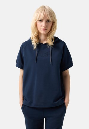 Blonde vrouw met een marineblauwe korte mouwen hoodie met tekst aan de voorkant, handen in zakken, kijkt vooruit op een effen lichte achtergrond.