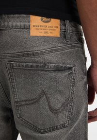 Blugi din denim gri cu un eticheta din piele maro cu logo, buzunare din spate cu cusături curbate și o suprafață de țesătură texturată.