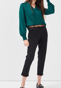Blouse col en V couleur sarcelle avec manches froncées, associée à un pantalon chino noir retroussé et une ceinture tressée beige. Des mocassins noirs complètent la tenue.