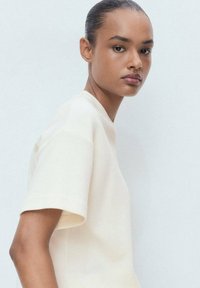 Top a maniche corte color crema realizzato in un morbido tessuto con un design pulito. Presenta una vestibilità rilassata e cuciture sottili lungo le spalle.