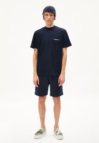 Marineblauw katoenen t-shirt met een zak, gecombineerd met bijpassende marineblauwe shorts. Schoeisel heeft een bloemmotief op een lichte zool.
