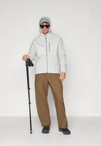 Veste gris clair avec capuche et fermeture éclair turquoise, pantalon cargo marron avec plusieurs poches, chaussures de randonnée noires, et un bâton de trekking.
