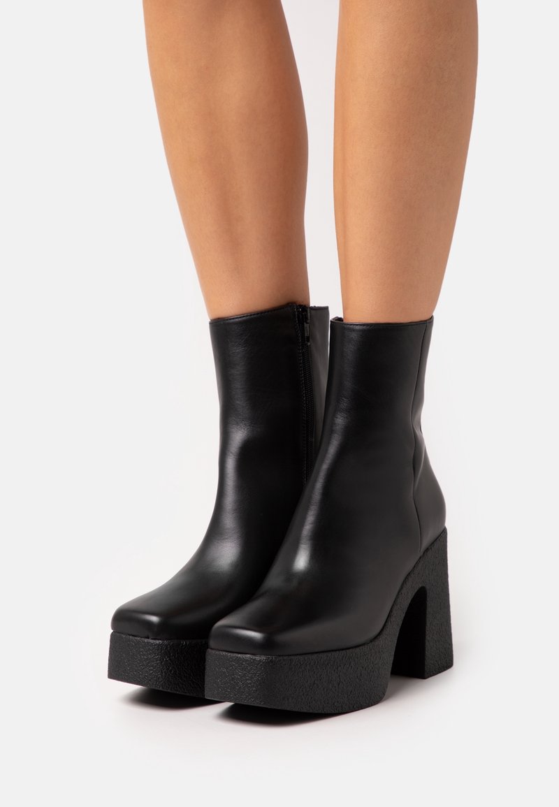 Bottines noires en cuir avec talons hauts et épais à plateforme, portées sur des jambes nues sur un fond clair uni.
